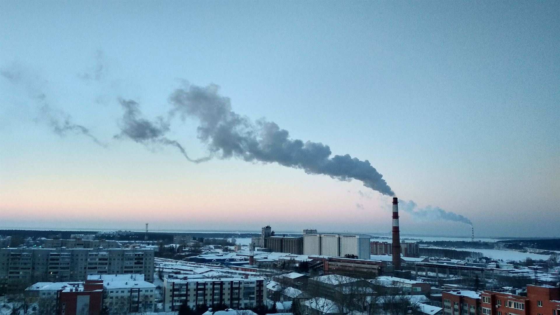 Шлейф дыма из промышленной трубы над заснеженным городом